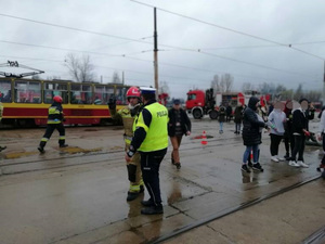 Uczestnicy podczas symulacji katastrofy. Policjant i strażak rozmawiają obok stoją statyści, w tle tramwaj i wóz straży  pożarnej.