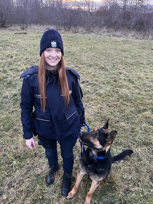 Policjantka w mundurze stojąca z psem służbowym. Pies patrzy się na funkcjonariuszkę.