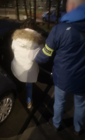 Policjant pomaga zatrzymanej kobiecie wsiąść do samochodu.