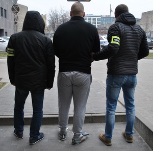 Policjanci prowadzą zatrzymanego, który ma założone kajdanki na nogi i ręce tak zwane zespolone.