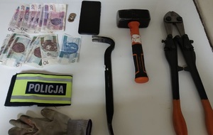 Narzędzia, telefon komórkowy i pieniądze zabezpieczone przy sprawcy leżą na stole, obok opaska z napisem policja.