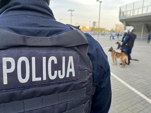 Policjant stoi przed stadionem w tle przewodnicy z psami służbowymi.