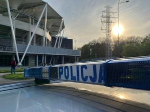 Belka z napisem policja na radiowozie w tle stadion.