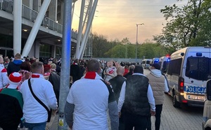 Grupy kibiców kierujące się w stronę wejścia na stadion. Obok po prawej stronie stoją oznakowane radiowozy.