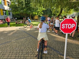 Dzieci na placu, dziewczynka na rowerze zaczyna jazdę po torze, obok znak drogowy stop.