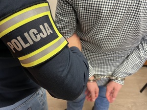 Policjant trzyma mężczyznę, który ma kajdanki założone na ręce trzymane z tyłu.