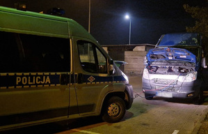 Oznakowany radiowóz policyjny typu bus z podpiętymi kablami rozruchowymi podłączponymi do drugiego samochodu.
