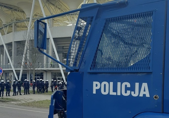 Policyjna armatka wodna pod stadionem, w tle policjanci zabezpieczający wydarzenie.