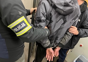Policjanci trzymają mężczyznę, który ma kajdanki założone na ręce trzymane z tyłu.
