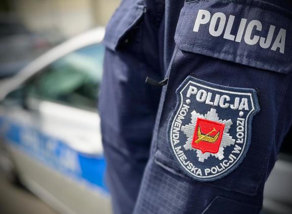 Naszywka Komendy Miejskiej Policji w Łodzi na ramieniu bluzy munduru, w tle oznakowany radiowóz.