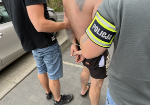 Policjanci trzymają mężczyznę, który ma kajdanki założone na ręce trzymane z tyłu.