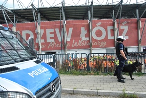 Oznakowany radiowóz policyjny oraz przewodnik z psem służbowym na tle stadionu Widzewa Łódź.