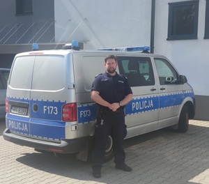 Umundurowany policjant stoi przed oznakowanym radiowozem.