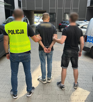 Mężczyzna z założonymi na ręce z tyłu kajdankami jest trzymany przez policjantów.