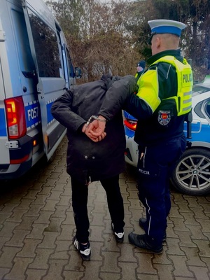 Mężczyzna z kajdankami założonymi na ręce trzymane z tyłu jest trzymany przez policjanta z łódzkiej drogówki.