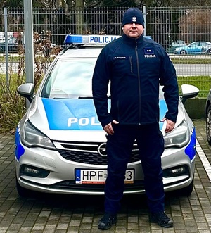 Umundurowany policjant stoi przed oznakowanym radiowozem policyjnym.