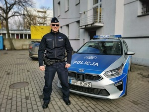 Umundurowany policjant stoi przed oznakowanym radiowozem.