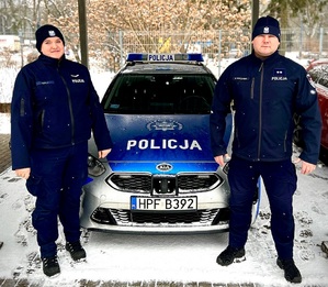 Umundurowani policjant i policjantka stoją przed oznakowanym radiowozem.