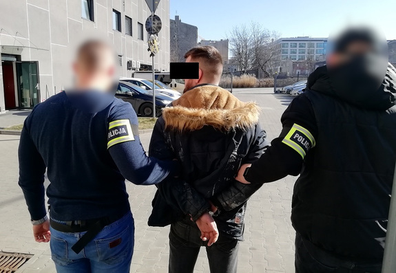 Policjanci trzymają mężczyznę, który ma kajdanki założone na ręce z tyłu.