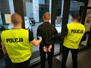 Policjanci trzymają mężczyznę, który ma kajdanki założone na ręce trzymane z tyłu.