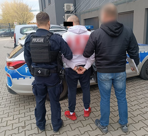 Policjanci trzymają mężczyznę, który ma kajdanki założone na ręce trzymane z tyłu.