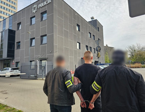 Policjanci trzymają mężczyznę, który ma kajdanki założone na ręce trzymane z tyłu.