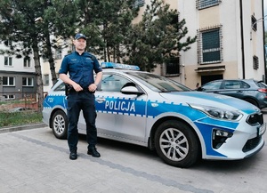 Umundurowany policjant stoi przed oznakowanym radiowozem.