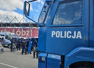 Na pierwszym planie policyjna armatka wodna w tle funkcjonariusze podczas zabezpieczenia spotkania piłkarskiego.