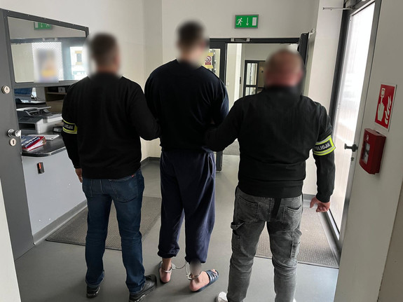 Policjanci z zatrzymanym mężczyzną, który ma założone kajdanki zespolone.