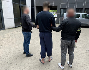 Policjanci prowadzą zatrzymanego mężczyznę, który ma założone zespolone kajdanki.
