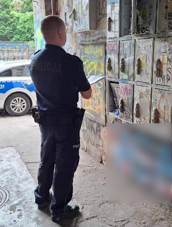 Dzielnicowy stoi przed ścianą z licznikami prądu.