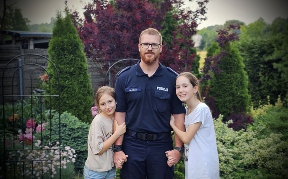 Do umundurowanego policjanta przytulają się dwie dziewczynki, jego córki.