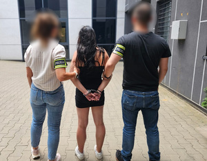 Policjantka i policjant trzymają kobietę, która ma kajdanki założone na ręce z tyłu.