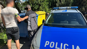 Policjant trzyma zatrzymanego mężczyznę, za nimi stoją kosze na śmieci, a obok nich oznakowany, policyjny radiowóz.