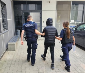 Policjanci prowadzą mężczyznę, który ma kajdanki zespolone założone na ręce i nogi.