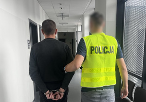Policjant trzyma mężczyznę, który ma kajdanki założone na ręce z tyłu.