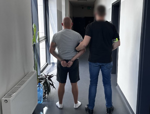 Policjant trzyma mężczyznę, który ma kajdanki założone na ręce z tyłu.