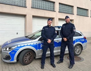 Umundurowani policjanci stoją przed oznakowanym radiowozem.