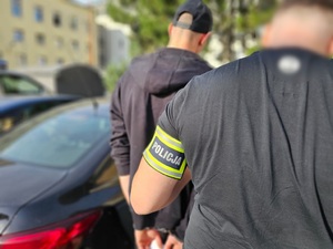 Policjant pomaga wsiąść do radiowozu mężczyźnie, który ma kajdanki założone na ręce trzymane z tyłu.