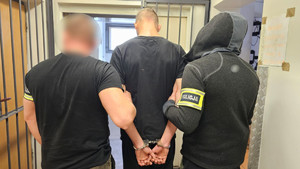 Policjanci trzymają mężczyznę, który ma kajdanki założone na ręce z tyłu.