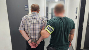 Policjant trzyma mężczyznę, który ma kajdanki założone na ręce z tyłu.