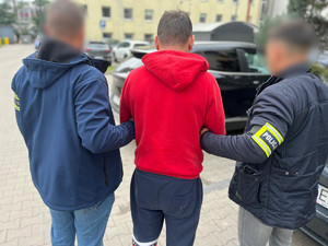 Policjanci trzymają mężczyznę, który ma założone kajdanki zespolone.