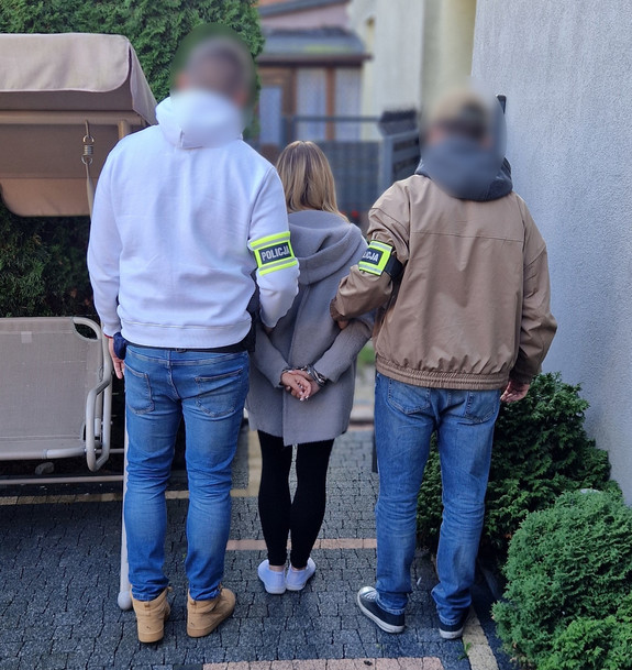Policjanci trzymają kobietę, która ma kajdanki założone na ręce z tyłu.