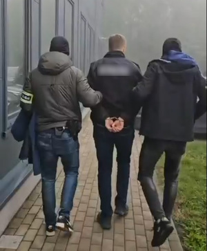 Policjanci prowadzą mężczyznę, który ma kajdanki założone na ręce z tyłu.
