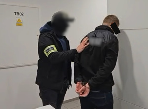 Policjant trzyma mężczyznę, który ma kajdanki założone na ręce z tyłu.