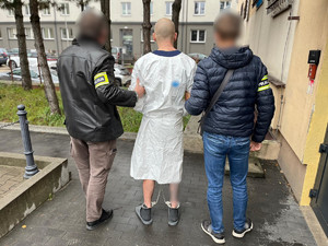 Policjanci trzymają mężczyznę, który ma założone kajdanki zespolone.