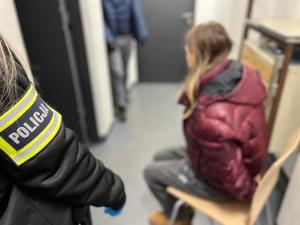 Opaska z napisem "Policja" na ramieniu, w tle, na krześle siedzi kobieta z kajdankami założonymi na ręce z tyłu.