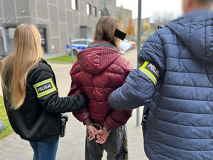 Policjanci trzymają kobietę, która ma założone kajdanki na ręce z tyłu.