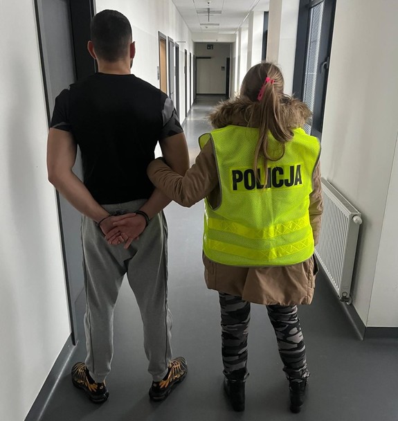 Policjantka trzyma mężczyznę, który ma kajdanki założone na ręce z tyłu.