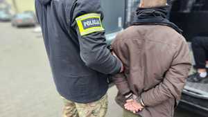 Policjant trzyma mężczyznę, który ma kajdanki założone na ręce z tyłu.